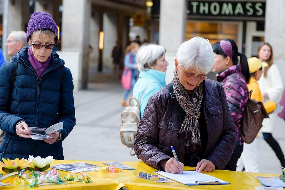 Žena potpisuje peticiju protiv progona Falun Gonga, dok druga prolaznica čita Falun Gong letak.