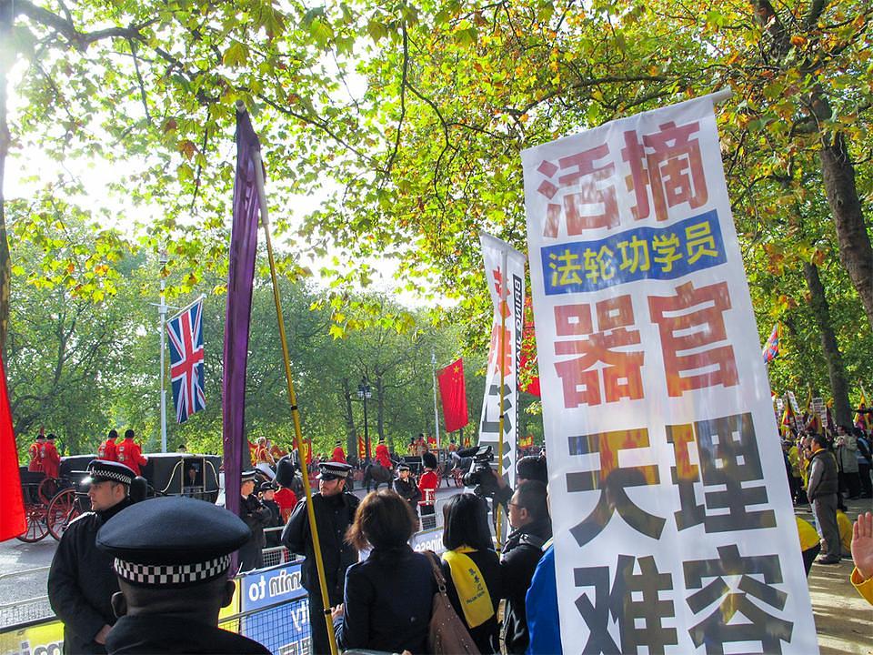 Pratnja predsjednika Xija prolazi pokraj Falun Gong transparenata. Na jednom transparentu sa slike piše: „Nebesa zabranjuju prisilnu žetvu organa od Falun Gong praktikanata“ 